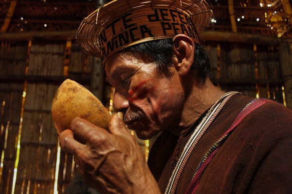 Benjamin Tiviito Coshanti, in a traditional hat and with red face paint, holds a bowl to his face to take a drink in a straw and wood building.