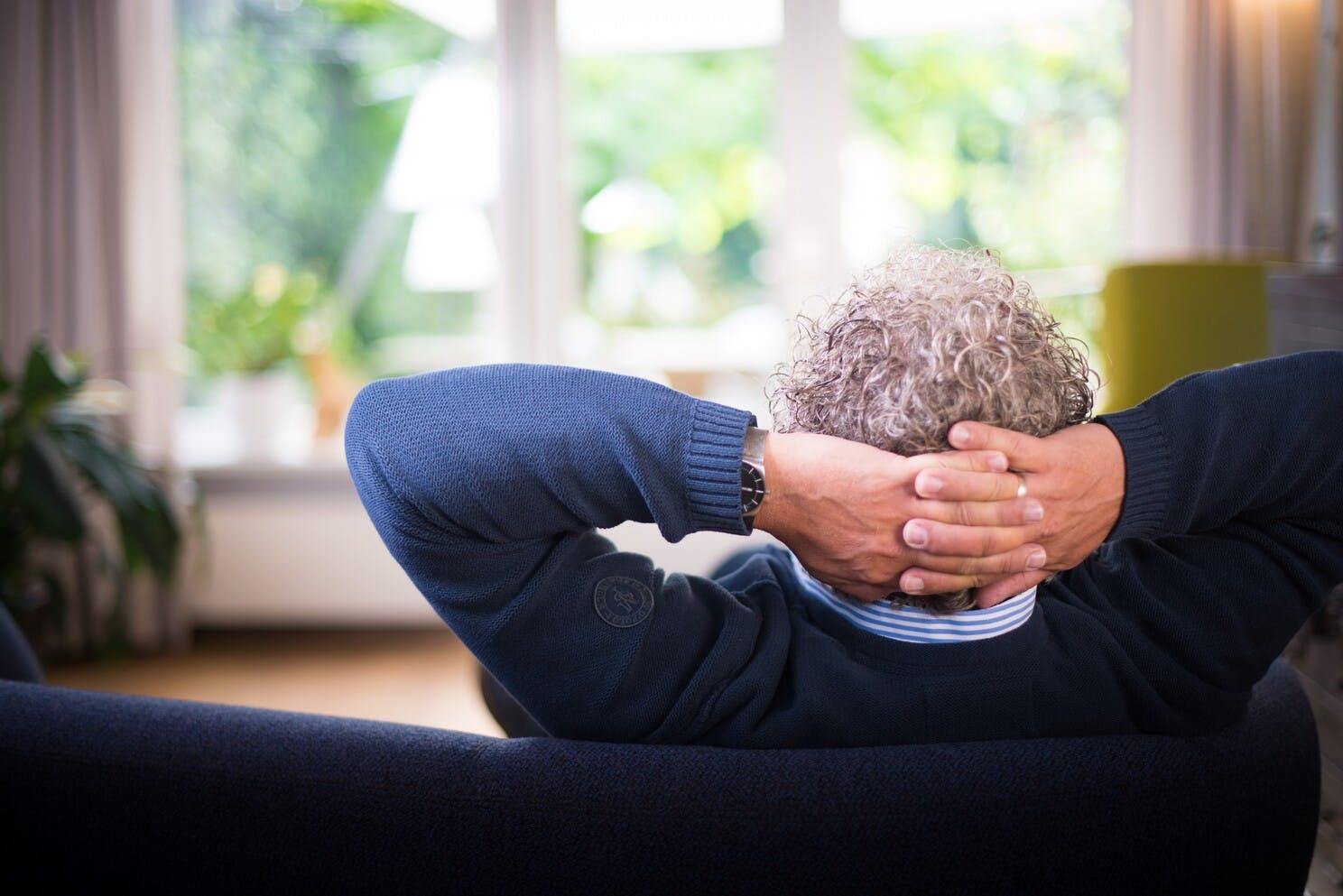 Un hombre mayor está comodamente recostado en un sofá viendo la hermosa vista del jardín por la ventana y con las manos en la nuca para descansar el cuello.